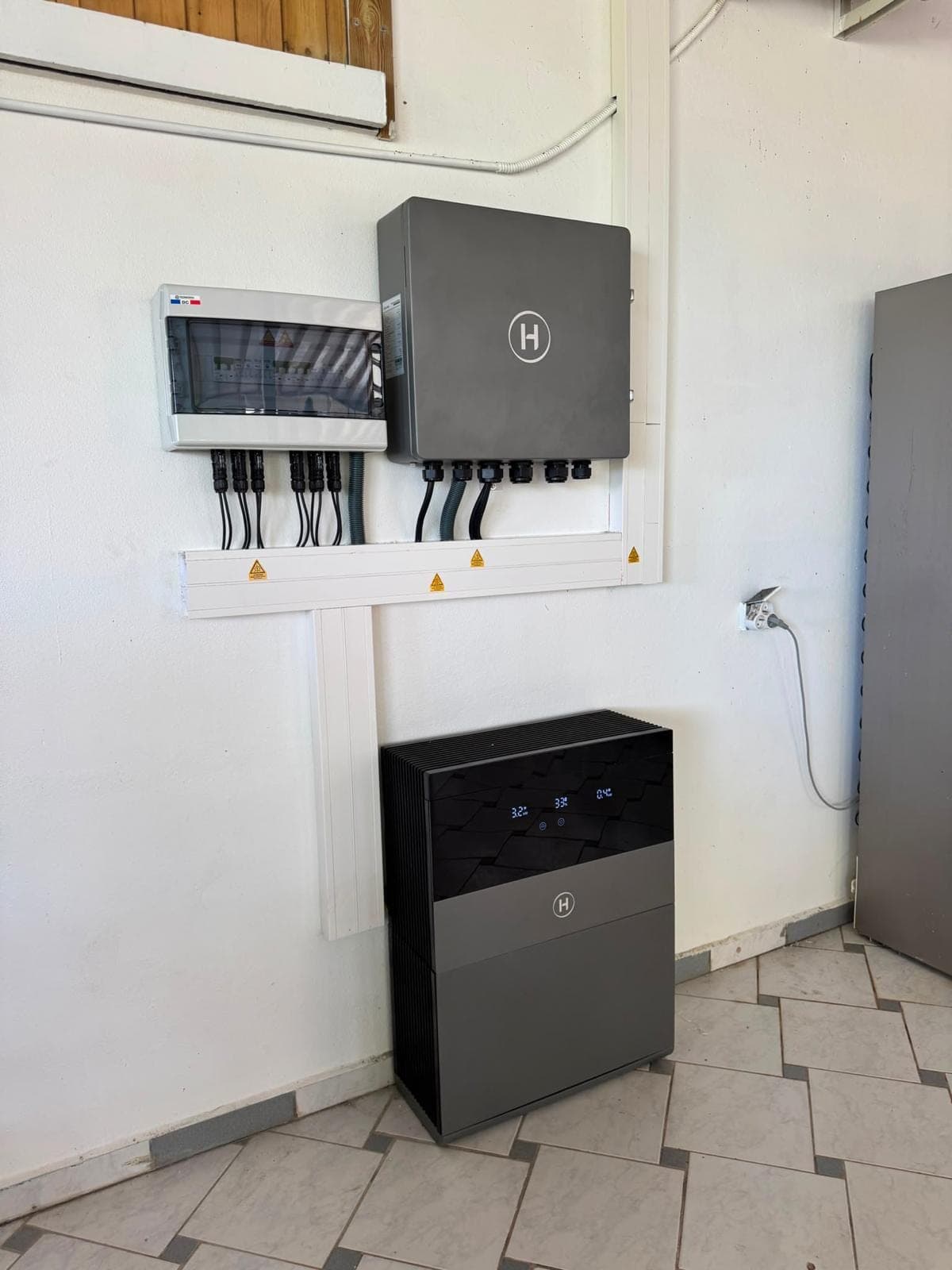 Onduleur hybride et batterie installés dans un local technique carrelé, coffrets de protection au mur